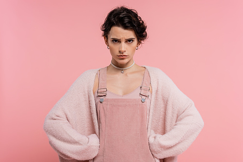 angry woman in strap dress and warm cardigan standing akimbo and looking at camera isolated on pink
