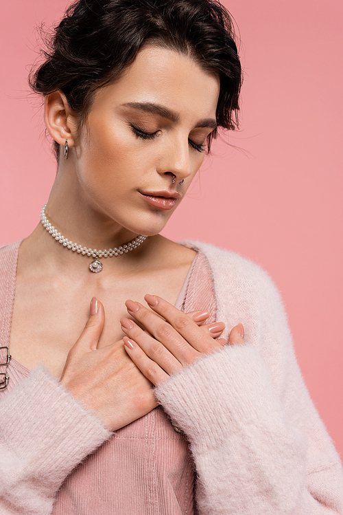 young woman in pearl necklace and piercing posing with hands on chest and closed eyes isolated on pink