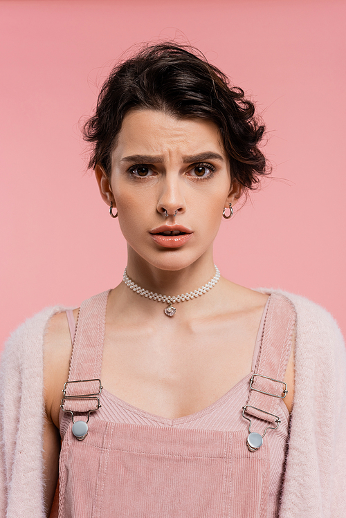 worried woman in strap dress and pearl necklace looking at camera isolated on pink