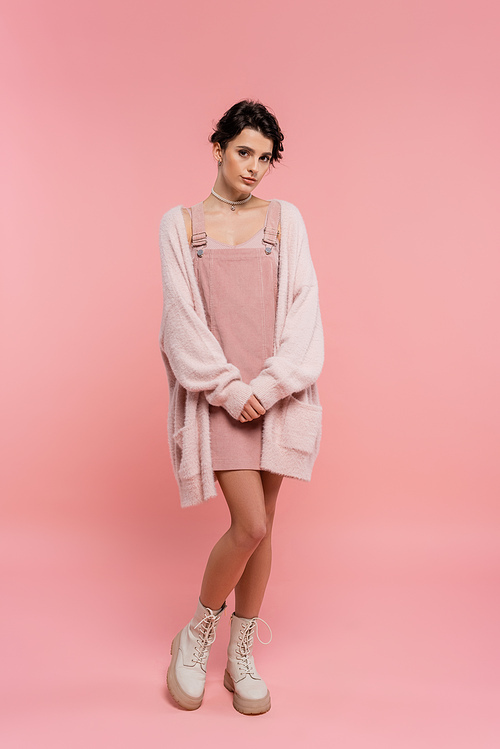 full length of brunette woman in boots and strap dress with warm cardigan looking at camera on pink background