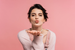 pretty brunette woman in cozy cardigan sending air kiss and looking at camera isolated on pink