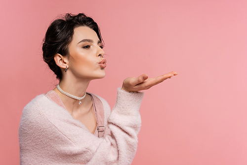 trendy woman in fluffy cardigan and pearl necklace sending air kiss while looking away isolated on pink