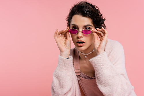 brunette woman in pearl necklace touching trendy sunglasses while looking at camera isolated on pink
