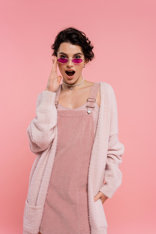 astonished woman in cozy cardigan adjusting trendy sunglasses while looking at camera isolated on pink
