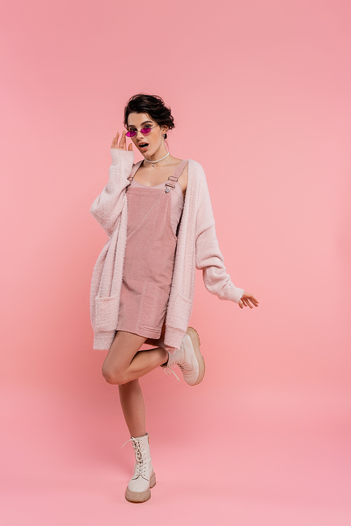 full length of amazed woman in sunglasses and fluffy cardigan posing on pink