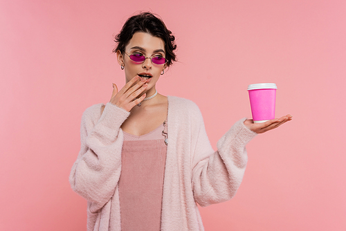 thoughtful woman in stylish sunglasses holding hand near mouth while looking at takeaway drink isolated on pink