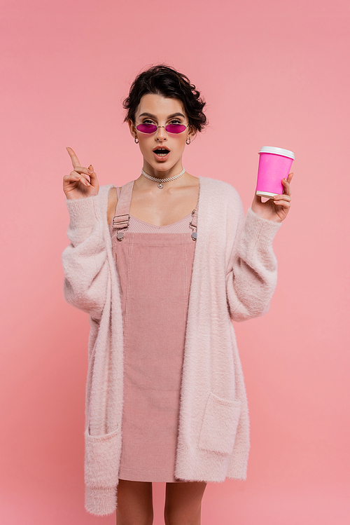 amazed woman in warm cardigan and sunglasses holding paper cup and pointing with finger isolated on pink