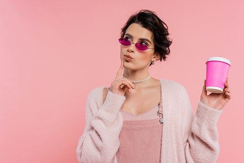 thoughtful woman in trendy sunglasses holding takeaway drink and looking away isolated on pink