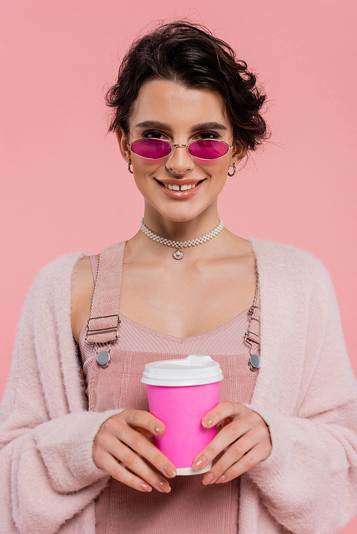 cheerful woman in stylish sunglasses and warm cardigan holding coffee to go isolated on pink