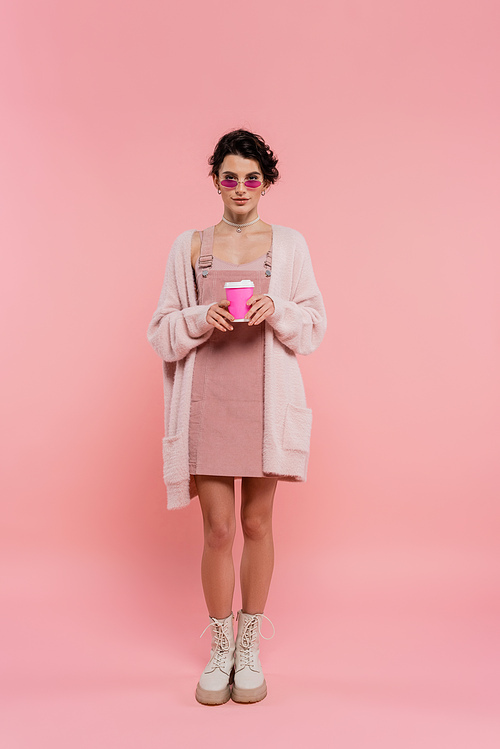 full length of woman in warm cardigan and trendy sunglasses holding coffee to go on pink