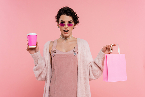 amazed woman in sunglasses and warm cardigan holding shopping bag and paper cup isolated on pink