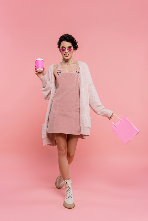 full length of fashionable woman walking with paper cup and shopping bag on pink
