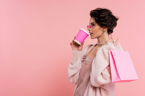 brunette woman in trendy sunglasses drinking coffee to go while holding shopping bag isolated on pink