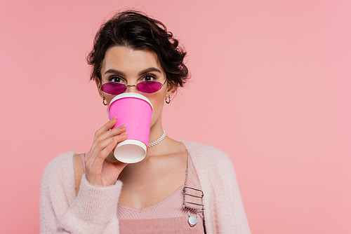 brunette woman in trendy sunglasses drinking coffee from paper cup isolated on pink