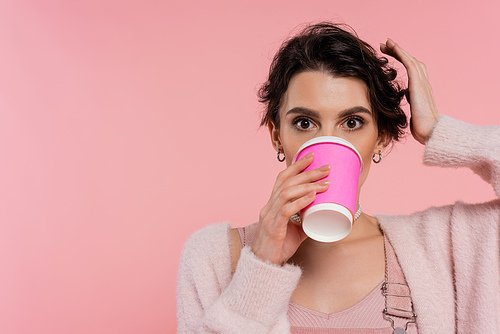 brunette woman looking at camera while drinking coffee from paper cup isolated on pink