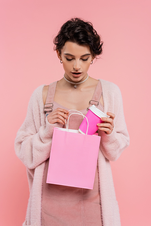 brunette woman in cozy cardigan putting paper cup into shopping bag isolated on pink