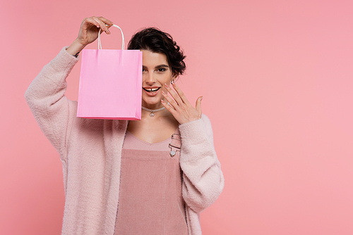 happy woman in soft cardigan showing shopping bag and holding hand near face isolated on pink