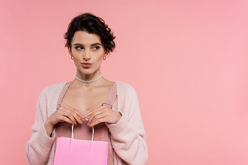 tricky brunette woman in warm cardigan holding shopping bag and looking away isolated on pink