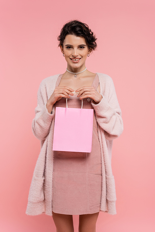 pleased brunette woman in warm cardigan holding shopping bag and smiling at camera isolated on pink