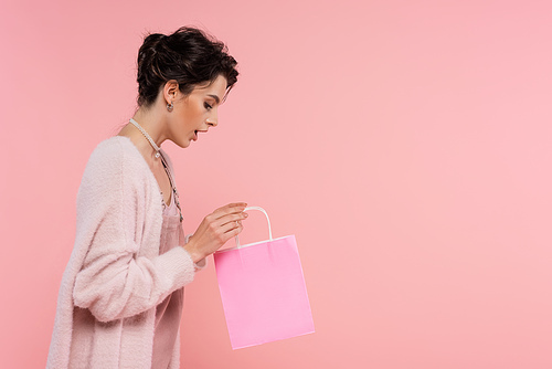 side view of amazed brunette woman in warm cardigan looking into shopping bag isolated on pink