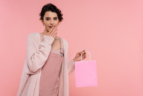 amazed woman in warm cardigan holding shopping bag and covering open mouth with hand isolated on pink