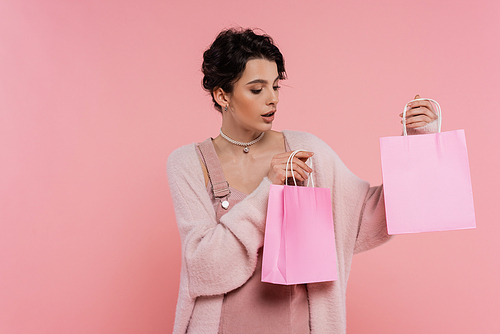 amazed young woman in fluffy cardigan holding shopping bags isolated on pink