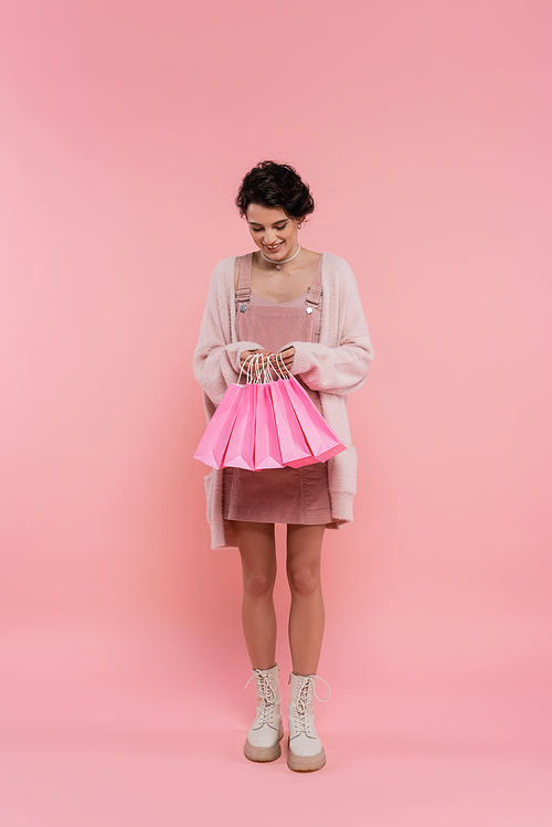 full length of pleased woman in warm cardigan and boots looking at shopping bags on pink background