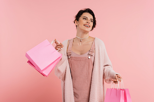 excited woman in strap dress and cozy cardigan holding shopping bags and looking away isolated on pink