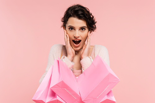 astonished woman with shopping bags touching face and looking at camera isolated on pink