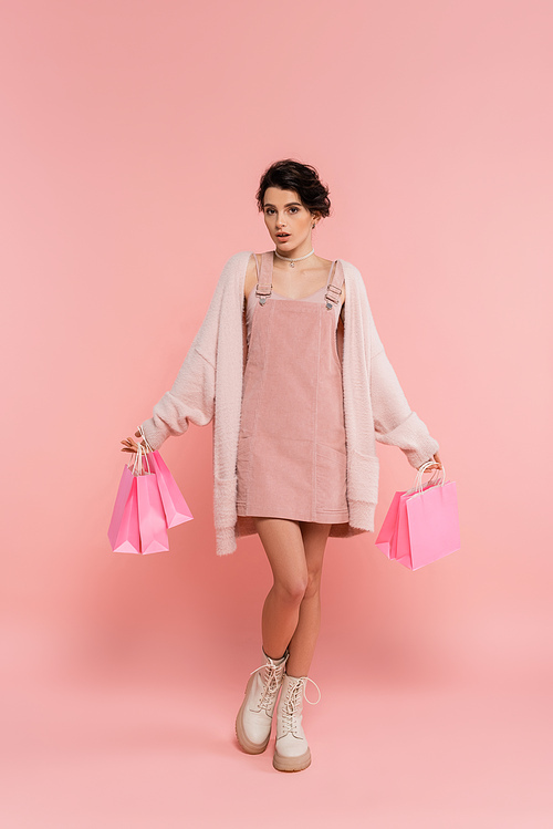 full length of trendy and surprised woman looking at camera while holding shopping bags on pink