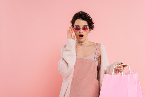 astonished woman with shopping bags touching trendy sunglasses and looking at camera isolated on pink