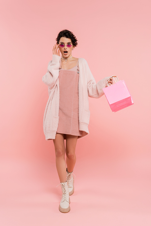 fashionable and surprised woman touching sunglasses while walking with shopping bags on pink