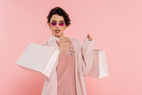 dreamy woman in trendy sunglasses holding shopping bags and looking away isolated on pink
