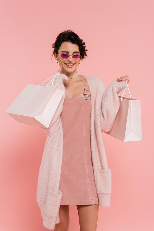 smiling woman in trendy sunglasses and warm cardigan posing with shopping bags isolated on pink
