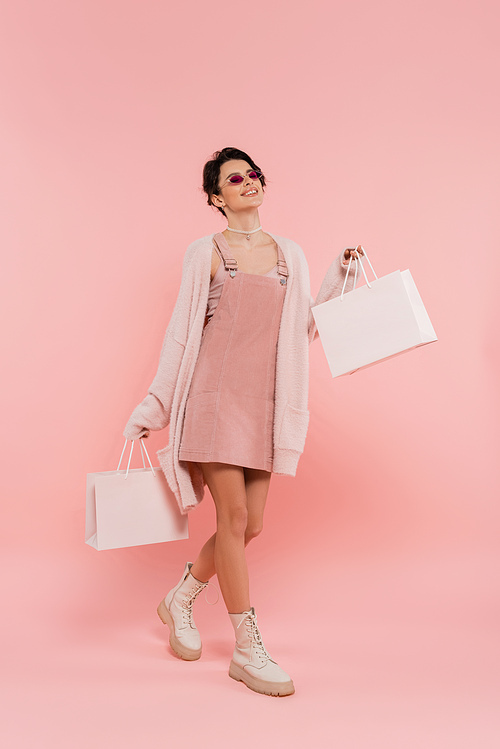 smiling woman in sunglasses and trendy clothing walking with shopping bags on pink