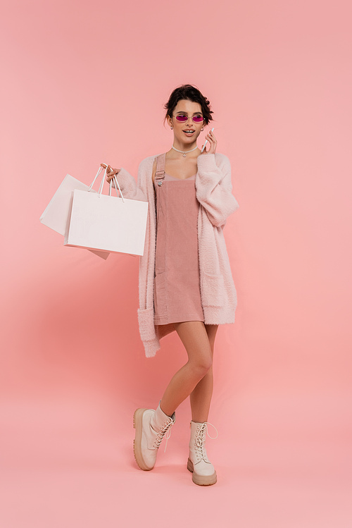 full length of woman in sunglasses and warm cardigan holding shopping bags and talking on smartphone on pink