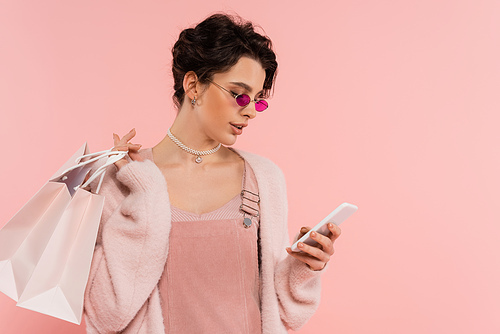 woman in trendy sunglasses holding shopping bags and messaging on smartphone isolated on pink