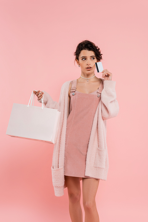 thoughtful brunette woman in warm cardigan holding shopping bag and credit card while looking away isolated on pink