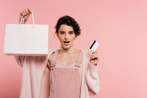 astonished woman with credit card and shopping bag looking at camera isolated on pink