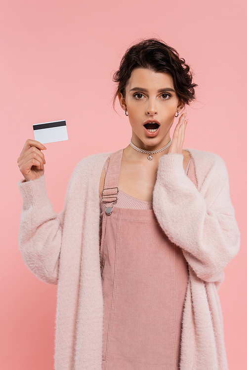 thrilled woman in warm cardigan holding credit card and hand near face isolated on pink