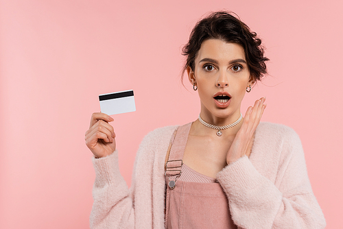 astonished woman holding hand near face while showing credit card isolated on pink