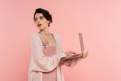 astonished brunette woman with laptop looking back isolated on pink
