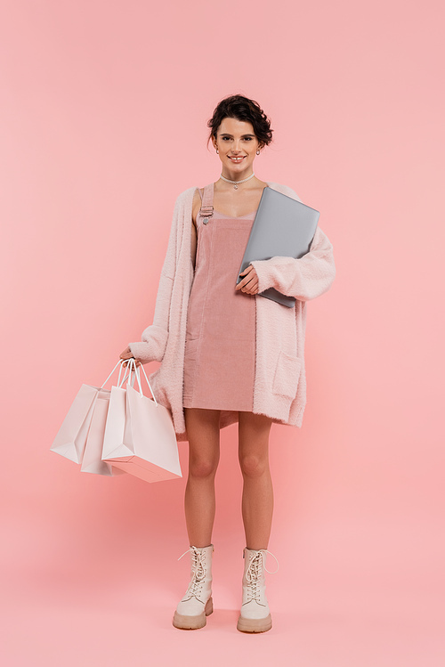 full length of trendy woman with laptop and shopping bags smiling at camera on pink background