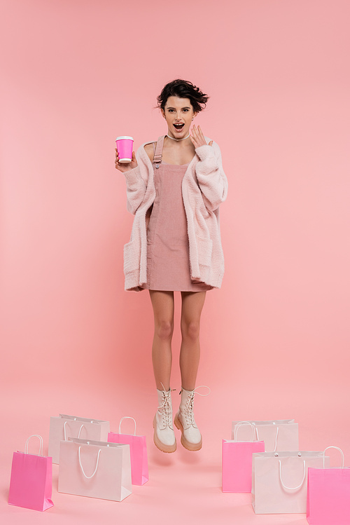 full length of astonished woman with coffee to go levitating near shopping bags on pink background