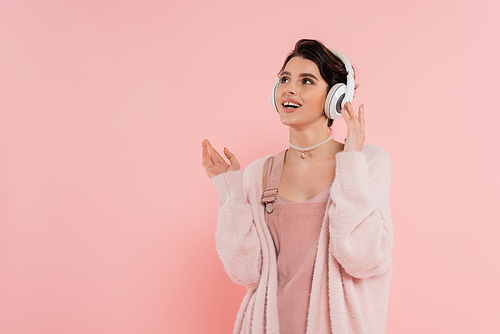 amazed and cheerful woman in wireless headphones looking away isolated on pink