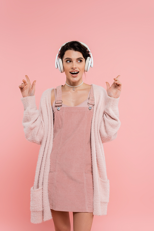 excited woman in strap dress and warm cardigan listening music in wireless headphones isolated on pink