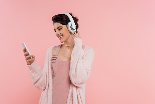 happy brunette woman in wireless headphones looking at mobile phone isolated on pink