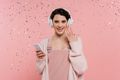 cheerful brunette woman in wireless headphones holding smartphone under confetti on pink background