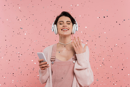 joyful woman with closed eyes holding cellphone and listening music in wireless headphones near confetti on pink