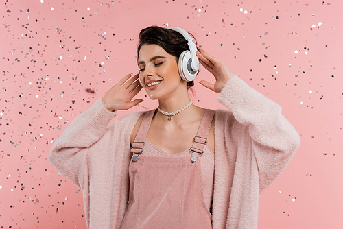 pleased woman touching wireless headphones while listening music under confetti on pink background